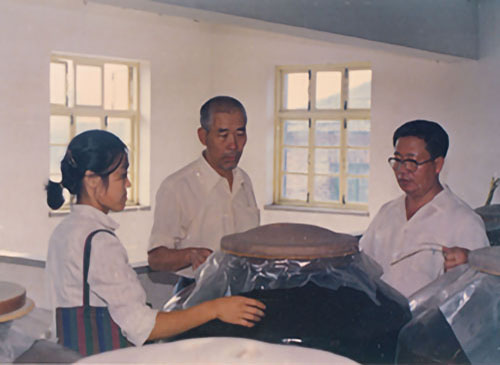 1992年，著名(míng)釀酒專家周恒剛（中(zhōng)）在景芝釀酒車(chē)間指導工(gōng)作(zuò)